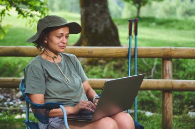 Doğayla iç içe olan bir kadın dışarıda dizüstü bilgisayar kullanarak oturuyor. Sahne, barışçıl, çevre dostu, harmanlama teknolojisi ve açık hava rahatlaması.