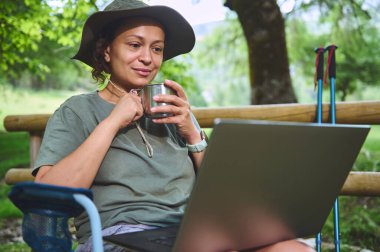 Bir kadın açık havada yemyeşil bir ortamda oturuyor, laptopuyla çalışırken sıcak bir içkinin tadını çıkarıyor. Sahne, boş zaman ve verimliliği doğal bir ortamda birleştiriyor..