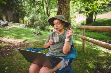 Ormandaki bir alanda oturan bir insan içkinin keyfini çıkarırken dizüstü bilgisayarla çalışıyor. Uzak çalışma, macera ve doğayla bağlantıyı temsil eder..