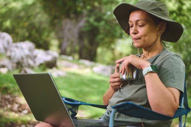 Doğal ortamında katlanabilir bir sandalyede oturan, elinde metal bir bardak tutan ve dizüstü bilgisayar kullanan, konfor, iş ve doğanın uyumunu somutlaştıran bir kadının sakin bir sahnesi..