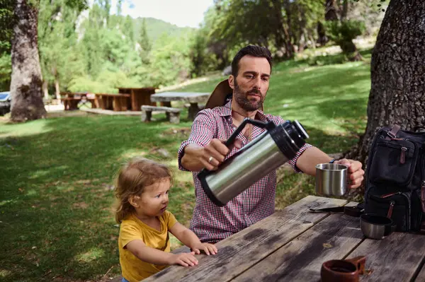 Bir baba ve genç kızı, yeşil bir ortamda, ahşap bir masada oturarak, doğanın tadını çıkarırken, kaliteli aile zamanlarının tadını çıkararak, dışarıda huzurlu bir ortam paylaşıyorlar..