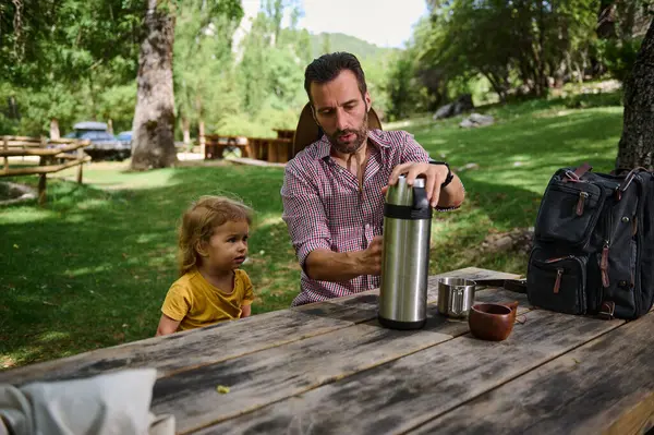 Küçük çocuklu bir adam dışarıda sakin bir anı paylaşıyor. Bereketli yeşilliklerle çevrelenmiş olarak, manzaralı bir günde kaliteli zaman geçirmekten hoşlanırlar..