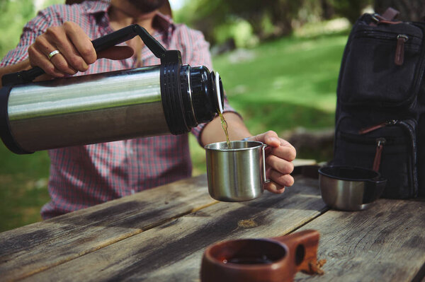 A person pours a warm drink into a metal mug from a thermos on a wooden table in a natural setting, representing outdoor leisure and relaxation in a serene environment.