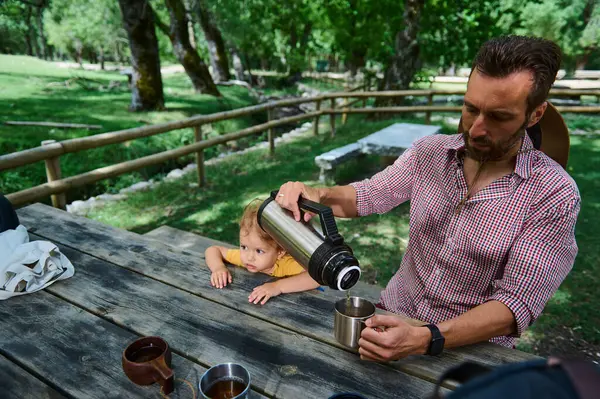 Güneşli bir günde sakin bir ormanda piknik yaparken bir adam termostan sıcak bir içecek döker..