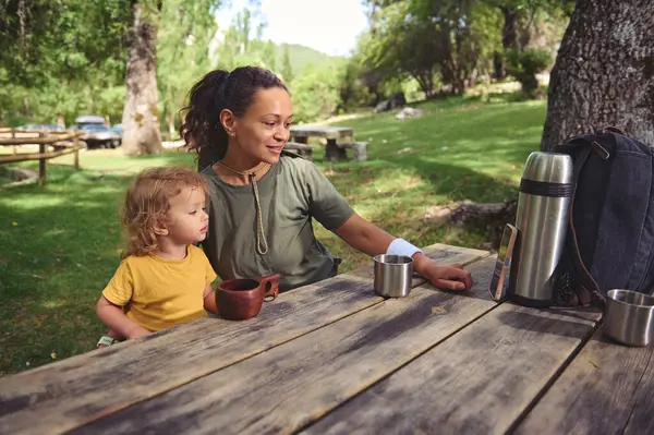 Bir anne ve çocuğu piknik masasında otururken sakin bir açık hava anında, doğayla bağlantıları teşvik ediyor ve sakin bir park ortamında sessiz-odaklanma aktivitelerini kucaklıyorlar..