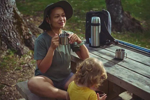 Bir anne, doğayla iç içe olan bir piknik bankında oturmuş çocuğuyla huzurlu bir anı paylaşıyor. Huzurlu bir yemek ve içeceğin tadını çıkarıyor..