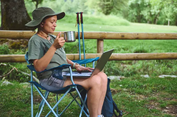 Açık havada sakin bir ortamın tadını çıkaran sıcak kahveyi yudumlarken dizüstü bilgisayarla çalışan biri. Göstergeler rahatlama, uzak çalışma ve doğanın ortasında bağlı bir yaşam tarzı.