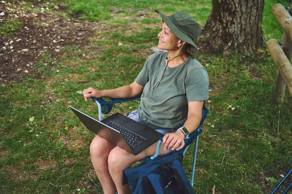 Bir kadın, açık hava ortamında bir kamp sandalyesine oturmuş dizüstü bilgisayarı ile gülümsüyor. İş ve boş zamanı birleştiriyor, uzak çalışma yaşam tarzını doğal ortamda somutlaştırıyor..