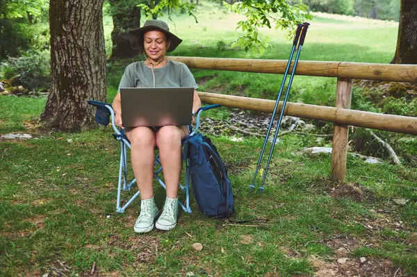 Dışarıda sakin bir yerde oturan bir kadın dizüstü bilgisayarı üzerinde çalışıyor. Doğal ortam ve yürüyüş ekipmanları onun doğa zevkini ve açık hava yaşam tarzını gösteriyor..