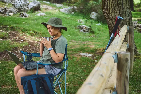 Yeşil şapkalı genç bir kadın, sakin bir açık hava ortamında dizüstü bilgisayarda çalışıyor. Doğal ortamda bağlantı ve verimlilik gösteriyor..