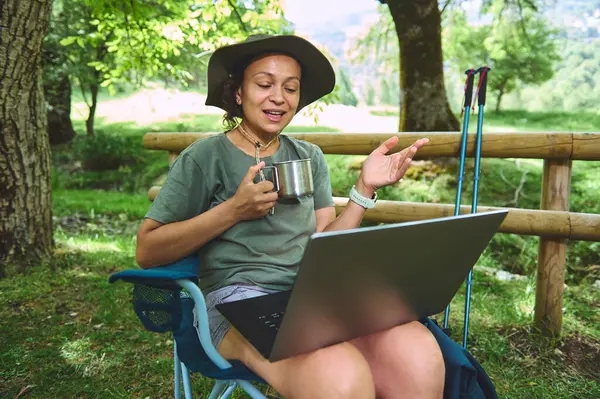 Bir kadın dijital göçebe yaşam tarzından hoşlanır, doğada dizüstü bilgisayar kullanır. Çevrimiçi etkileşimde, içkisini yudumluyor ve yemyeşil bir yaşam tarzını kucaklıyor..