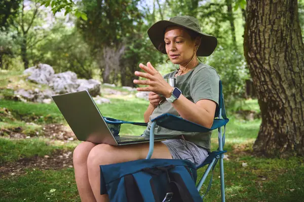 Katlanan bir sandalyede oturan bir kadın, bir dizüstü bilgisayarla çalışırken aynı zamanda ormanın temiz havasının ve huzurunun tadını çıkarıyor, doğanın ve dijital göçebenin özünü somutlaştırıyor..