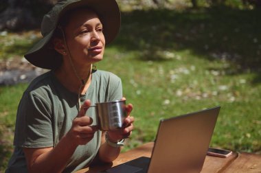 Kadın dışarıda uzaktan çalışıyor, laptopunu kullanarak bir kupayla ferahlatıcı bir mola veriyor, doğal manzarayla çevrili dengeli bir yaşam tarzını somutlaştırıyor..