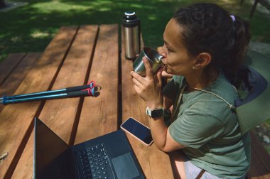 Bir kadın laptopuyla dışarıda çalışmaktan zevk alıyor, bir içecek yudumluyor, dijital göçebeliği ve doğadaki uzak çalışma hayatını temsil ediyor..