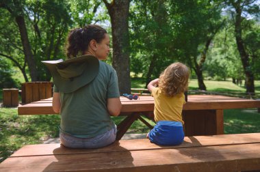 Bir anne ve çocuğu yemyeşil bir parktaki piknik masasında birlikte oturuyorlar. Dış mekan doğal güzellik ve sükuneti gösteriyor. Bağlanma ve gevşeme anlarını yansıtıyor..