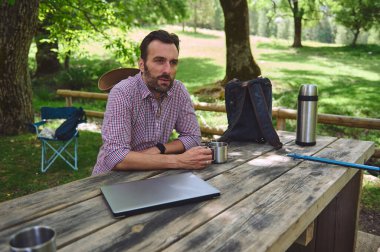 Yeşil ormandaki ahşap bir piknik masasında, elinde metal bir bardak, dizüstü bilgisayar, termos ve sırt çantasıyla rahatlayan bir adam dijital göçebe yaşam tarzını ve boş zamanlarını temsil ediyor..