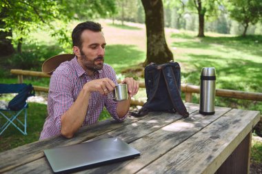 Bir adam kahve içerken güneşli bir parkta ahşap bir masada oturuyor. Bilgisayarı, sırt çantası ve termosu dijital göçebe yaşam tarzını gösteriyor..