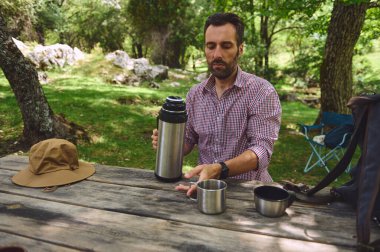 Sıcak bir şeyler içerken doğanın tadını çıkaran bir adam. Canlı yeşilliklerle çevrili bir piknik masasında, huzurlu ve rahat bir atmosfer sergiliyor..