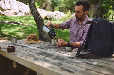 Huzurlu bir orman ortamının ortasında ahşap bir masada otururken bir adam termostan bardağa kahve dolduruyor. Güneş şapkası ve sırt çantası Macera katıyor.