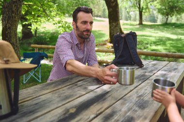 Kırsal bir ahşap piknik masasında oturan bir adam manzaralı bir ortamda karşıdaki birine metal bardak sunuyor. Bağlantının vurgulandığı rahat bir açık hava sahnesi.