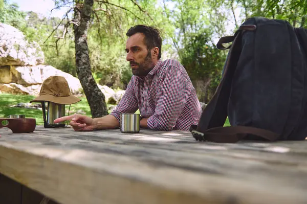 Doğada oturan bir adam, çevresi yeşilliklerle çevrili, tahta bir masaya kamp malzemeleri yerleştirilmiş, sakin bir kamp ortamını ve muhtemelen dijital göçebe yaşam tarzını somutlaştırıyor..