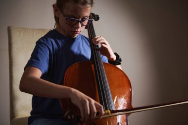 Genç müzisyen yumuşak ışıkta çello çalışmasına odaklanmış. Huzurlu bir ortamda müzikal yetenek ve adanmışlık gösterisi..