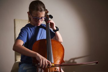Gözlüklü genç bir adam, bir sandalyeye oturmuş, stüdyoda ustaca çello çalıyor, sıcak bir ışık ve odaklanmış bir ifadeyle..