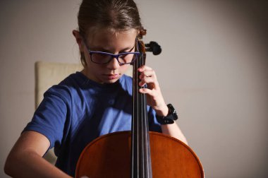 Gözlüklü genç bir kız dikkatle çello çalıyor, odaklandığını ve kendini müziğe adadığını gösteriyor. Dekor, onun müzik ve pratik yapma konusundaki ciddi çalışmasını tamamlıyor..