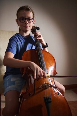 Genç bir çello sanatçısı, günlük kıyafetlerini evde, odaklanmış aydınlatma altında giyer ve sakin bir ortamda müziğe olan bağlılıklarını ve yeteneklerini vurgular..