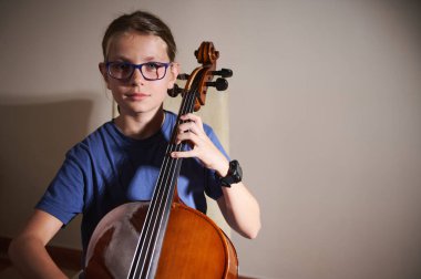 Sıradan giyinmiş, evde oturmuş, ustalıkla çello çalan genç bir adam. Işık yumuşak gölgeler yaratıyor, sahnenin dingin ve odaklanmış atmosferini vurguluyor..