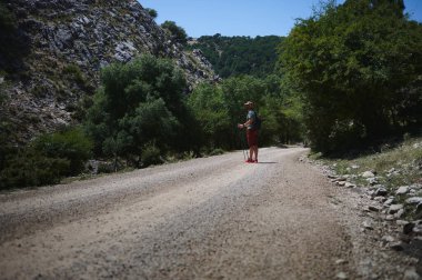 Yeşillik ve kayalık tepelerle çevrili toprak bir yolda yürüyen bir yürüyüşçü. Doğada macera ve keşifler..