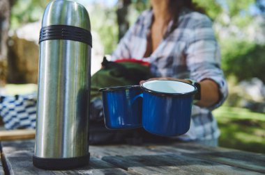 Mavi diş mineleri ve piknik masasına yerleştirilmiş metal termoslu açık hava seti. Arka planda, sakin bir orman ortamında kamp malzemeleri organize eden biri var..