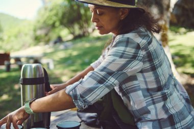 Günlük kıyafet giymiş bir kadın güneşli bir parkta piknik masasında oturup içki hazırlıyor. Manzaralı ortam ve onun barışçıl aktivitesi eğlenceli bir açık hava deneyimini gösteriyor..