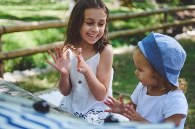 Açık havada güneşli bir günün tadını çıkaran iki genç kız. Parkta gülümseyip el çırpıyorlar. Atmosfer mutlulukla ve çocukluk etkileşimiyle doludur..