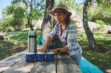 Güneşli bir günde, açık hava rahatlığı ve rahatlığı yansıtan, canlı yeşilliklerle çevrili ahşap bir masada bir kadının fincanlar organize ettiği sakin bir piknik sahnesi..
