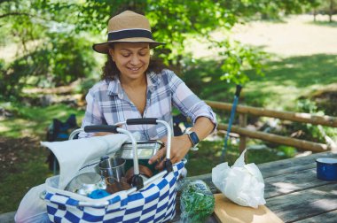 Bir kadın yemyeşil bir parkta eğlenirken piknik sepeti düzenliyor. Ortam sakin ve rahatlamak, doğanın tadını çıkarmak ve dışarıda kaliteli zaman geçirmek için ideal..