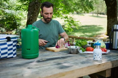 Bir adam açık hava pikniği sırasında bir yemek hazırlarken sebze dilimler. Huzurlu doğal ortam, açık hava yemeklerini ve sağlıklı yaşamı ön plana çıkaran sakin bir arka plan sağlar..