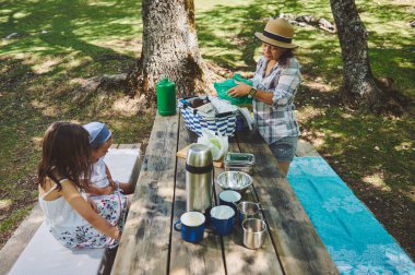 Çocuklarıyla birlikte bir anne, doğayla ve yeşilliklerle çevrili güneşli bir günde açık havada piknik yapmaktan hoşlanır. Sahne huzurlu ve neşeli bir aile aktivitesini tasvir ediyor..