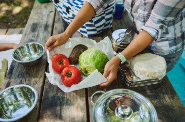 Açık hava yemeği sırasında paslanmaz çelik kaplarla çevrili kırsal bir piknik masasında marul, domates ve avokado da dahil olmak üzere taze sebzeleri düzenleyen eller..