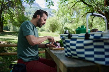 Bir adam açık havada ahşap bir masada oturuyor, manzaralı bir orman ortamında piknik için sebze dilimliyor. Çizgili bir sepet ve yeşil termos yazın sakin atmosferini güçlendirir..