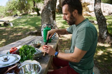 Bir adam dışarıda piknik masasında oturuyor, yemek için sebze hazırlıyor. Ayar güneşli bir günde yeşilliklerle çevrili sakin bir park..