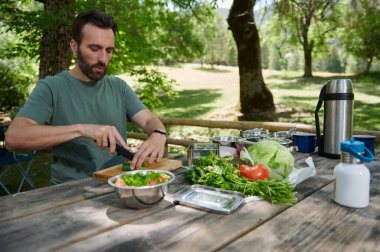 Yeşillikte, ahşap bir piknik masasında taze salata malzemeleri hazırlayan bir adam. Sağlıklı malzemeler ve doğal ışık ile mükemmel bir açık hava yemek sahnesi..