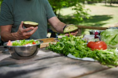 Avokado yarım tutan bir kişi, açık hava ortamında ahşap bir masada taze bir salata hazırlarken. Taze sebze ve sebzeler sağlıklı ve doğal bir yemek önerir..