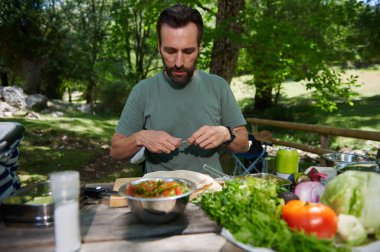 Bir adam taze sebzelerle sağlıklı bir yemek hazırlarken dışarıda huzurlu bir pikniğin tadını çıkarır. Doğanın ortasında geçen sahne, dingin doğal ortamların rahatlamak için mükemmel olduğunu vurgular..