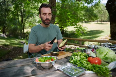 Bir adam, açık havada, canlı sebzelerle çevrili ahşap bir masada taze bir salata hazırlarken bir avokadoyu dilimliyor ve doğal ve rahat bir ortam yaratıyor..