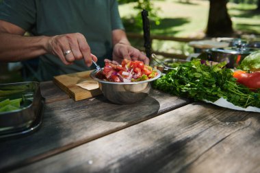Elleri açık hava pikniği sırasında taze yeşillik ve malzemelerle çevrili canlı bir sebze salatasını doğal gün ışığında bir kasede karıştırırken görülüyor..