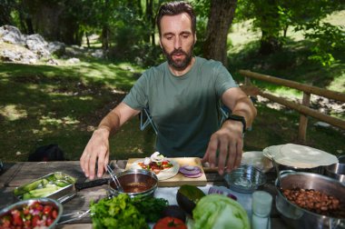 Bir adam açık havada, yemyeşil ağaçlar ve güneş ışığıyla çevrili taze bir yemek yapar. Doğal bir kaçamağın tadını çıkarır. Sağlıklı ve dengeli beslenir..
