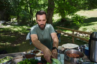 Yeşilliklerle çevrili bereketli, güneşli bir ormanda açık havada piknik masasında taze ürünler düzenleyen bir adam..