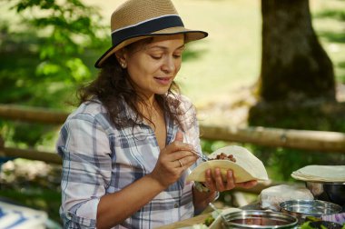 Tahta bir masada oturan bir kadın, sıradan bir şapka takıyor ve sakin bir doğal ortamda tortillayı fasulyeyle dolduruyor, boş vakitlerin tadını çıkarıyor..