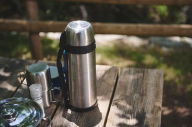 Güneşli bir günde, açık hava piknik masasında parlak çelik bir termos, fincanlar ve tuzluk. Orman arka planı dingin bir atmosfer ekler.